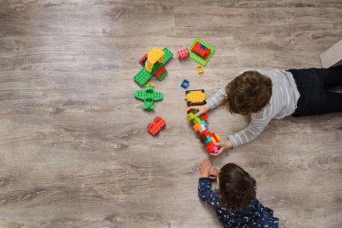 İki çocuk abisi ve kız kardeşi plastik oyuncak inşa tuğlalarıyla oynuyor. Ahşap zeminde yatıyorlar.