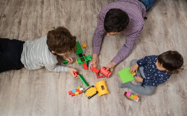 Babası ve iki çocuğu, erkek ve kız kardeşi plastik oyuncak tuğlalarla oynuyorlar. Ahşap zeminde yatıyorlar.