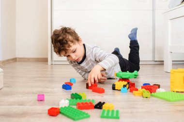 Küçük çocuk tahta zeminde yatıyor ve oyuncak plastik tuğlalarla oynuyor.