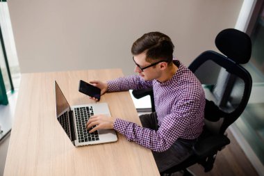 Yaklaş genç yakışıklı, rahat gömlekli, elinde akıllı telefon olan ve ofiste defter üzerinde çalışan adam..