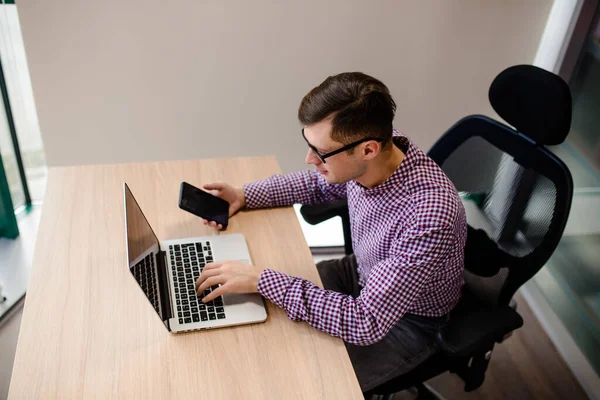 Yaklaş genç yakışıklı, rahat gömlekli, elinde akıllı telefon olan ve ofiste defter üzerinde çalışan adam..