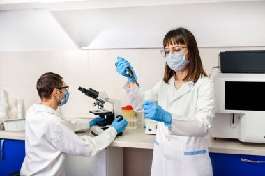 Hastane laboratuarlarında çalışan genç bir kadın doktor mikropipet test tüpleri kullanıyor. Arka planda laboratuvar asistanı immünolojik kimya için tam otomatik bir teşhis sisteminin yanındaki tıbbi mikroskoba bakıyor.