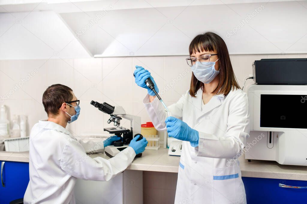 In the hospital laboratories working process, a young woman doctor is using micropipette test tubes, in the background a laboratory assistant is looking at the medical microscope near a fully-automated diagnostic chemistry system for immunological te