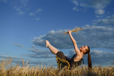 Beyaz noktalı siyah elbiseli mutlu genç kadın eski bir sandalyede oturuyor ve buğday tarlasında buğday tutuyor..