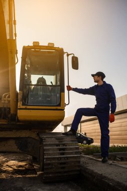Şantiyede gün batımı. Bir müteahhit silueti buldozerin yanında oturuyor. İnşaat makineleri konsepti