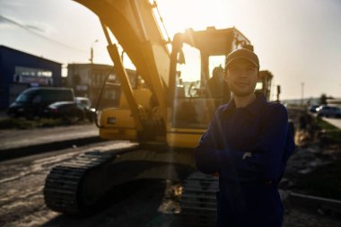 Mavi tulumlu gülümseyen müteahhit kameraya bakıyor. İnşaat makinelerinin bulunduğu arkaplan