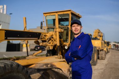 Mavi tulumlu gülümseyen müteahhit kameraya bakıyor. İnşaat makinelerinin bulunduğu arkaplan