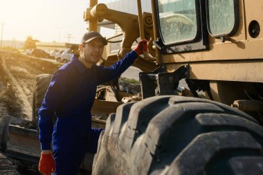 Mavi tulumlu gülümseyen müteahhit kameraya bakıyor. İnşaat makinelerinin bulunduğu arkaplan