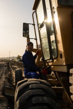 Mavi tulumlu genç inşaatçı inşaat alanındaki kazıcı makine parçalarına bakıyor.