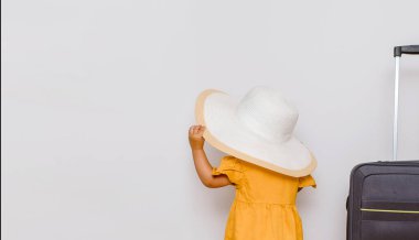 Büyük bir bavulun yanında beyaz şapkalı küçük bir kızın arka görüntüsü. Çocuklar bir tatil konsepti istiyor. Beyaz arkaplan kopyalama alanı