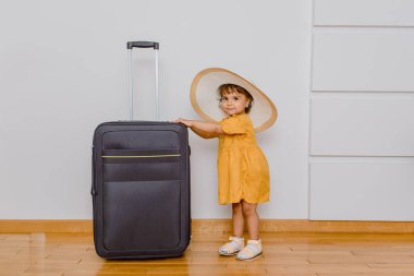 Büyük bir bavulun yanında beyaz şapkalı küçük bir kız. Çocuklar bir tatil konsepti istiyor.
