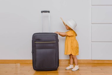 Büyük bir bavulun yanında beyaz şapkalı küçük bir kız. Çocuklar bir tatil konsepti istiyor.