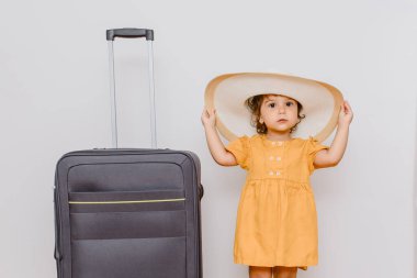 Büyük bir bavulun yanında beyaz şapkalı küçük bir kız. Çocuklar bir tatil konsepti istiyor..