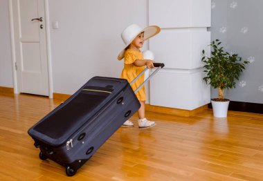 Büyük şapkalı ve sarı elbiseli küçük mutlu bir kız beyaz duvarları ve ahşap zemini olan büyük bir bavul taşıyor ve büyük bir tatil bekliyor.