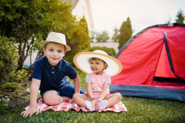 İki mutlu küçük çocuk, erkek ve kız kardeş ve kız kardeş evlerinin bahçesindeki kırmızı bir kamp çadırının yakınında çimlerde oturup oynuyorlar. Ev kampı tatil konsepti