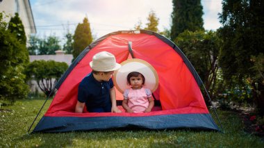 İki mutlu çocuk, erkek ve kız kardeş ve kız kardeş evlerinin bahçesindeki çimlerin üzerinde kırmızı bir kamp çadırında oynuyorlar. Ev kampı tatil konsepti
