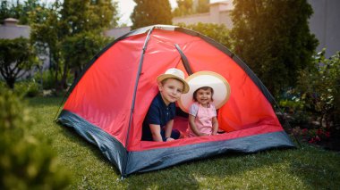 İki mutlu çocuk, erkek ve kız kardeş ve kız kardeş evlerinin bahçesindeki çimlerin üzerinde kırmızı bir kamp çadırında oynuyorlar. Ev kampı tatil konsepti