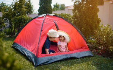 İki mutlu çocuk, erkek ve kız kardeş ve kız kardeş evlerinin bahçesindeki çimlerin üzerinde kırmızı bir kamp çadırında oynuyorlar. Ev kampı tatil konsepti