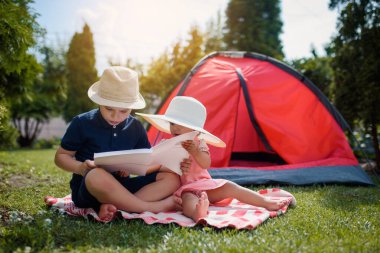 İki mutlu çocuk, erkek ve kız, erkek ve kız kardeş ve kız kardeş çimlerin üzerinde oturuyorlar ve erkek kardeş, evlerinin bahçesindeki kırmızı bir kamp çadırının yanında kız kardeşine peri masalları okuyor. Ev kampı tatil konsepti