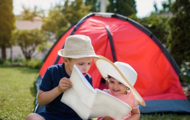 İki mutlu çocuk, erkek ve kız, erkek ve kız kardeş ve kız kardeş çimlerin üzerinde oturuyorlar ve erkek kardeş, evlerinin bahçesindeki kırmızı bir kamp çadırının yanında kız kardeşine peri masalları okuyor. Ev kampı tatil konsepti