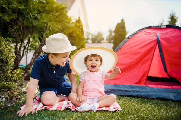 İki mutlu küçük çocuk, erkek ve kız kardeş ve kız kardeş evlerinin bahçesindeki kırmızı bir kamp çadırının yakınında çimlerde oturup oynuyorlar. Ev kampı tatil konsepti