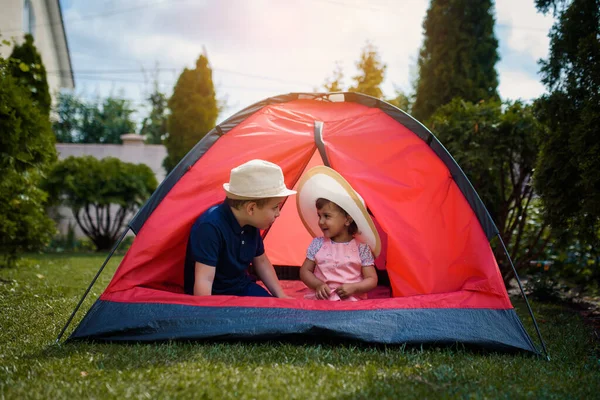 İki mutlu çocuk, erkek ve kız kardeş ve kız kardeş evlerinin bahçesindeki çimlerin üzerinde kırmızı bir kamp çadırında oynuyorlar. Ev kampı tatil konsepti