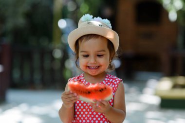 Kırmızı elbiseli, beyaz benekli ve şapkalı mutlu küçük kız çocuğu çok komik bir karpuz dilimi yiyor. Çocukluk kavramı komiktir.