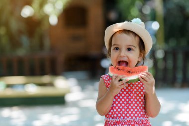 Kırmızı elbiseli, beyaz benekli ve şapkalı mutlu küçük kız çocuğu çok komik bir karpuz dilimi yiyor. Çocukluk kavramı komiktir.