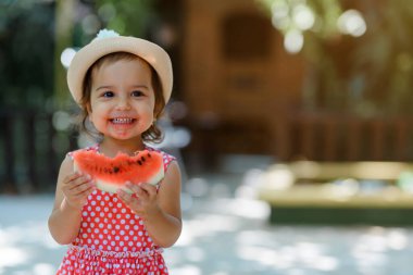 Kırmızı elbiseli, beyaz benekli ve şapkalı mutlu küçük kız çocuğu çok komik bir karpuz dilimi yiyor. Çocukluk kavramı komiktir.