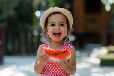 Kırmızı elbiseli, beyaz benekli ve şapkalı mutlu küçük kız çocuğu çok komik bir karpuz dilimi yiyor. Çocukluk kavramı komiktir.