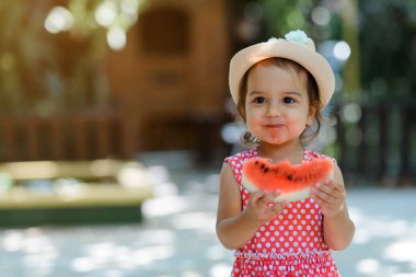 Kırmızı elbiseli, beyaz benekli ve şapkalı mutlu küçük kız çocuğu çok komik bir karpuz dilimi yiyor. Çocukluk kavramı komiktir.