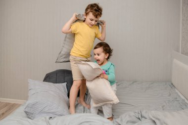 Mutlu aile oyunları konsepti. Küçük kız ve erkek kardeş yatakta yastıklarla oynuyorlar. Eğleniyorlar, gülüyorlar, gülüyorlar.