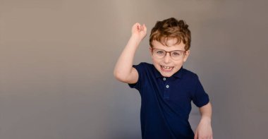 Mavi tişörtlü ve gri arka planda gözlüklü küçük çocuk parmağını göstererek gülüyor. Metin ve logo için boş alan