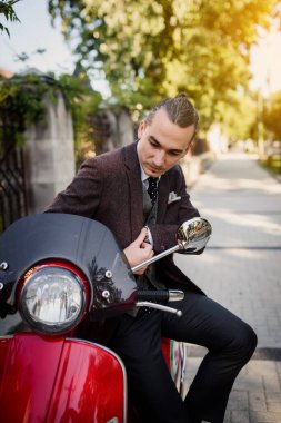 Zarif klasik ceketli ve yelekli genç yakışıklı adam kırmızı klasik motosiklet scooter 'ının yanında duruyor.