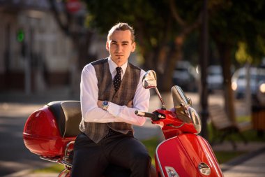 Beyaz tişörtlü, yakışıklı, klasik yelek ve kravatlı genç adam kırmızı klasik motosiklet scooter 'ında oturuyor ve kameraya bakıyor.