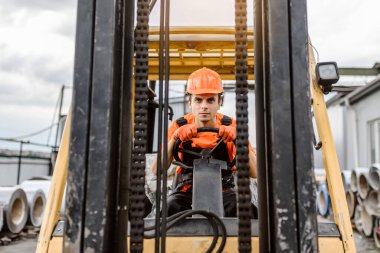 Ciddi bir yüzün ön görüntüsü. Tulumlu ve turuncu korumalı miğferli Kafkasyalı genç adam inşaat malzemeleri üzerine bir yük makinası sürüyor.