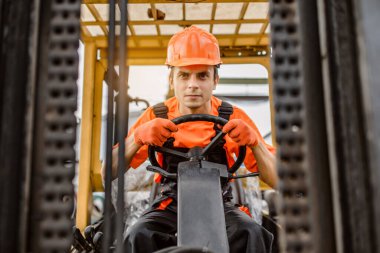 Ciddi bir yüzün ön görüntüsü. Tulumlu ve turuncu korumalı miğferli Kafkasyalı genç adam inşaat malzemeleri üzerine bir yük makinası sürüyor.