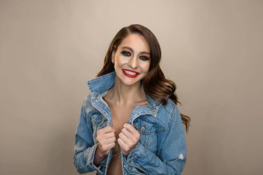 Mutlu gülümseyen genç modelin yakın portresi. Kırmızı dudaklı ve mükemmel ciltli. Gri arka planda kot ceket giyiyor.