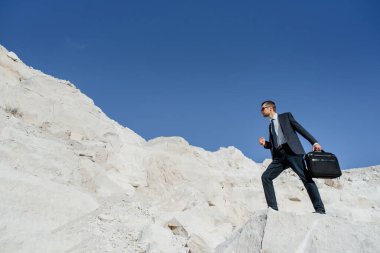 Çantalı ve ceketli genç iş adamı dağa tırmanıyor. İş hayatında başarı ve sıkı çalışma kavramı.