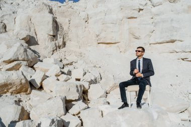 Klasik kıyafetler, ceket, kravat ve gözlüklü genç adam sandalyede oturuyor ve dağda poz veriyor..