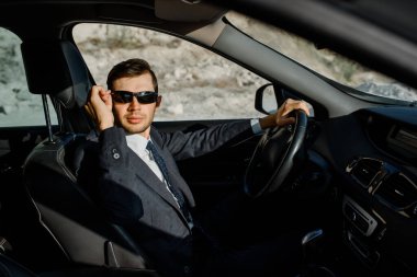 Ceketli, gömlekli, kravatlı ve güneş gözlüklü genç işadamı araba kullanıyor.