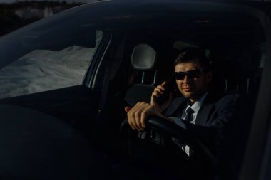 Ceketli, gömlekli, kravatlı ve güneş gözlüklü genç işadamı araba sürüyor ve akıllı telefondan konuşuyor.. 