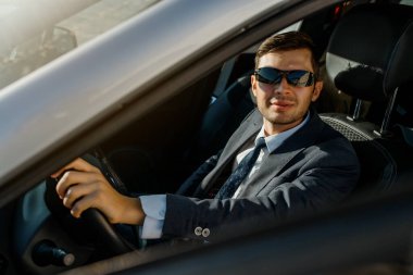 Ceketli, gömlekli, kravatlı ve güneş gözlüklü genç işadamı araba kullanıyor.