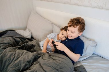 Mutlu erkek ve kız kardeşler gülümsüyor, gülüyor, yatakta uzanıyor, akıllı telefonda oynuyor ve eğleniyorlar.