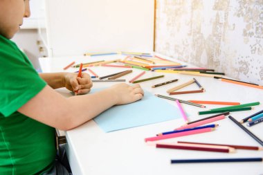 Mavi bir kağıda resim yapan bir çocuğun yan görüntüsü. Masanın üzerinde bir sürü renkli kalem kalemi var. Mutlu çocukluk kavramı