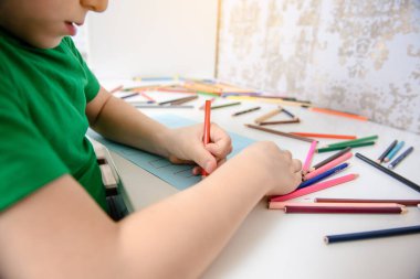 Mavi bir kağıda resim yapan bir çocuğun yan görüntüsü. Masanın üzerinde bir sürü renkli kalem kalemi var. Mutlu çocukluk kavramı