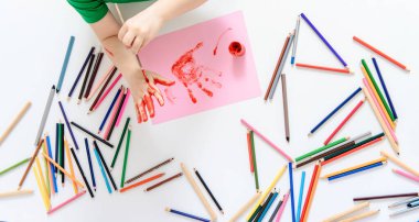  Elini kırmızı boyayla boyayan bir çocuğun en iyi görüntüsü. Bir sürü renkli kalem kalemi olan bir arka plan. Mutlu çocukluk kavramı