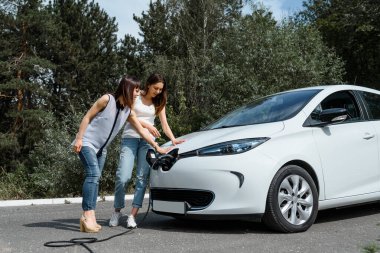 İki mutlu genç kadın, elektrikli arabaları ormandaki şarj istasyonunda şarj olana kadar birbirleriyle konuşuyorlar. Çevre dostu ulaşım konsepti