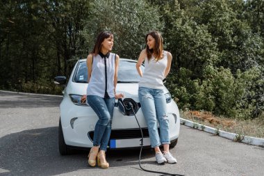 İki mutlu genç kadın, elektrikli arabaları ormandaki şarj istasyonunda şarj olana kadar birbirleriyle konuşuyorlar. Çevre dostu ulaşım konsepti