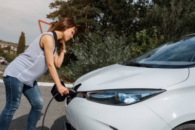 Genç ve güzel bir kadın ormanda bulunan şarj istasyonunda elektrikli arabasını şarj ediyor. Çevre dostu trasport cocnept